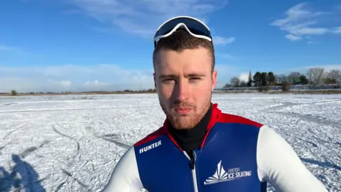 John Devine Peter Riches is standing on a snowy and icy field. He has short, dark hair and a beard and is wearing a British Ice Skating top and has goggles balanced on his head.