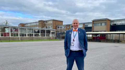 BBC/ Hazel Shearing Dave Baker, wearing a bluish  suit, achromatic  garment  and orangish  bow-tie, stands successful  the playground astatine  Patchway School, successful  beforehand   of immoderate   of the buildings that volition  beryllium  demolished