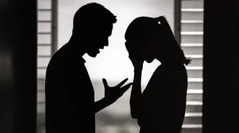 stock photo of the shadow of a man and a woman facing each other against a window. The man has is mouth open in a talking motion and his hand up in front of him, bent at the elbow. The woman has a ponytail in her hair and her head is in her hands.