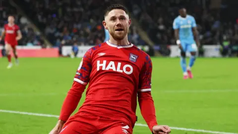 Getty Images A football player celebrates after a goal by sliding on the pitch. He is wearing a red football kit. 