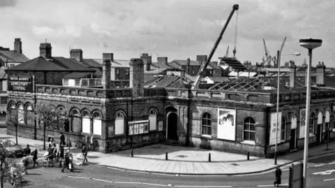 David Pearce A black and white image showing a single-storey Victorian railway station in Lowestoft. A crane is sticking out of the building removing its roof and its rafters are showing. On the left are men and women crossing a road. 
