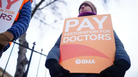 PA Media A person holding an orange BMA sign which says 'Pay restoration for doctors'. They are wearing a blue coat, pink scarf and pink hat. They are stood in front of a black iron fence which has a tree in the background. 