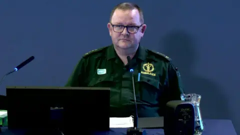 A man is sitting in front of him with a computer. He is wearing a paramedic uniform that calls an ambulance on him. It is a bottled green color. There is a jug of water on the table in front of him.