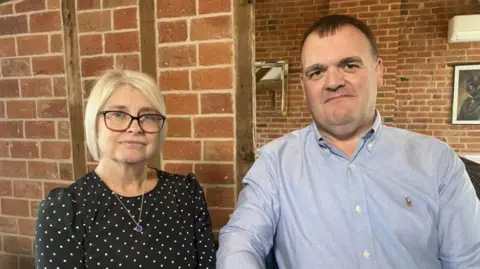A woman with blonde hair, glasses and a black top with white dots next to a man with short dark hair and a pale blue short, both standing in front of a brick wall