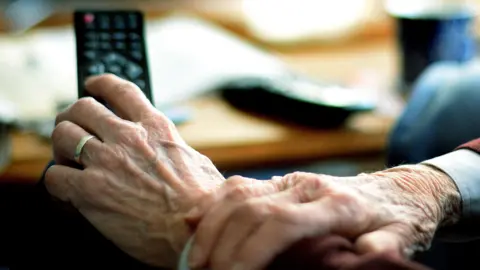 An elderly person watching TV. They are holding a TV remote in one of their hands.