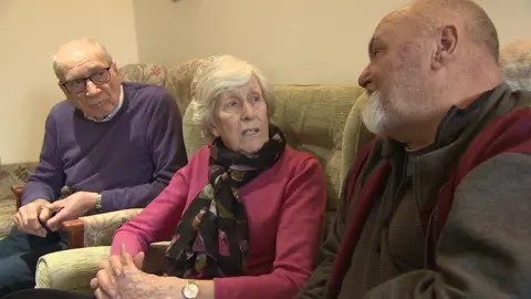 Three people are sitting in armchairs talking to each other in the communal lounge. Dorothy is sitting between her husband on her right and her neighbour Philip on her left. 