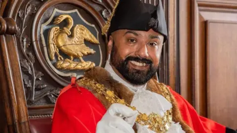A close-up image of Imran Hussain seated in an ornate wooden chair. He is dressed in a bright red robe trimmed with fur and black ceremonial hat with gold detailing.
