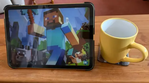 Martin Heath/BBC An iPad showing a scene from Minecraft, featuring a character wearing blue clothing fashioned from blocks. The iPad is standing on a table next to a yellow mug.