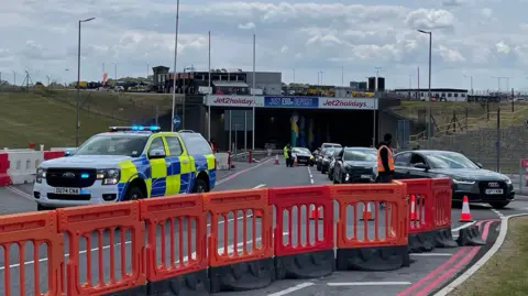 Helen Mulroy/BBC A police truck has its blue lights on behind a line of orange barriers shutting the road. It is blocking the entrance to a tunnel at the airport, which has a stream of cars queuing to get out.