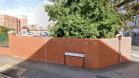 A red brick wall in front of a car park and a tree in Swindon. There is a white street sign with black lettering on in front of the wall, which reads "Edgeware Road". The sun is shining. There are buildings in the car park and down a street to the side.