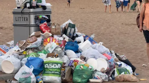 Silver waste bin with 'litter' written on it and litter overflowing on top and around it. It is on a sandy beach. There are people in the background.
