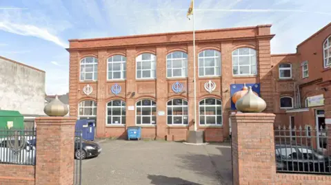 A rectangular brick building with large windows and colourful decorations on the wall. There is a car park with a flag pole in front of it and a gateway with two decorated brick posts