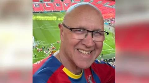 Rob Best. He is pictured standing in the stalls at Wembley Stadium. The football pitch can be seen in the distance behind him. Rob is wearing a football shirt and reading glasses. He is looking directly at the camera and smiling