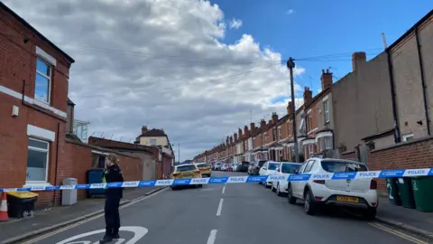 Street view of Newcombe Road. There is police tape blocking off entrance to the street and a police officer stood in the road.