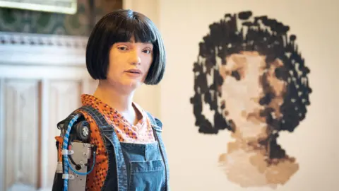 A robot that looks like a female with short dark hair looks towards the camera, she looks human but you can see the electronics in her arm.