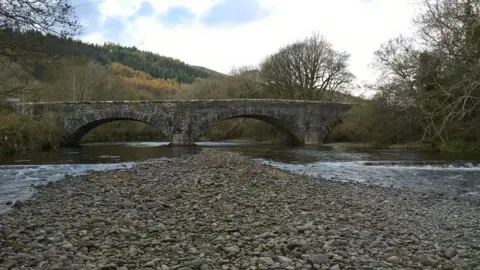 Westmorland and Furness Council Duddon Bridge pictured from a dry patch in the river. It is a stone bridge with three spans. The wide river is flowing and bubbling passed a central "island" of stones and pebbles. Trees overhang on each bank and beyond the bridge a hill rises, covered in dense woodland.