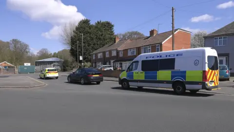 The photo shows a residential street scene with a marked police van and police car parked near terraced houses on a sunny day. Several civilian vehicles are also visible on the road, with trees and buildings in the background.