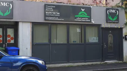 The street entrance to the mosque which has four windows and a door, with three signs saying IMC above them