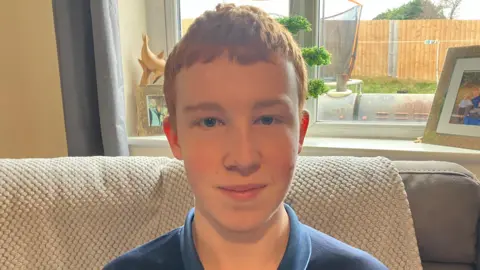 Teenager with red hair and blue eyes smiling at the camera sitting on a sofa in his home 