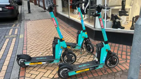Turquoise coloured e-scooters - there are three of them - parked on a pavement outside a clothes shop on a paved street.