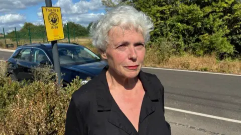A woman with short white hair stands beside a road near a blue car, with a yellow “Neighbourhood Watch Area” sign on a pole behind her and bushes and fencing in the background.