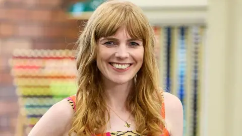 Close up of Adele, who has shoulder-length auburn hair stands in a brightly lit haberdashery area in the Sewing Bee studio