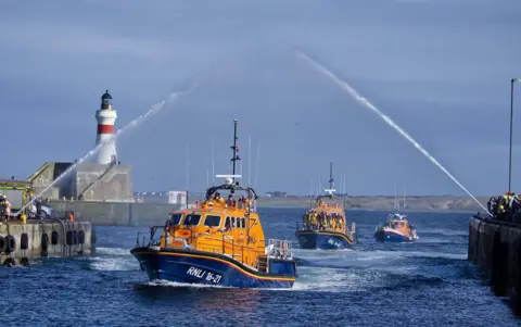 Mark Grant A parade of three orange RNLI lifeboats sailing into the harbour. There are crowds along either side of the entrance and a water arch is over the parade.