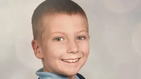 Watson family A young boy, Adam Watson, looks into the camera in what appears to be a school photograph. There is a plain background behind him and he is smiling broadly. He has very short brown hair.