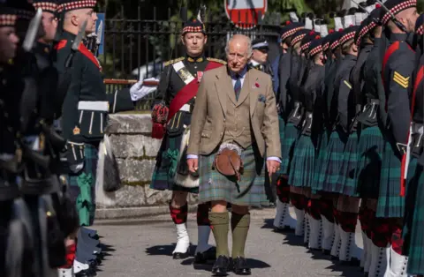 PA Media King Charles III, wearing a tweed jacket, waistcoat and tartan kilt, walks between two ranks of troops from Balaklava Company, 5th Battalion, The Royal Regiment of Scotland, during a ceremonial inspection. Soldiers in dark uniforms, tartan kilts and glengarry caps stand at attention on either side, creating a guard of honour along the roadway, while an officer in full ceremonial dress follows behind. The scene takes place outdoors in bright sunlight.