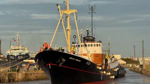 A sidewinder trawler which says Arctic Corsair on the side makes its way through a dock at sunrise. Several people can be seen on the trawler and by the side of it.