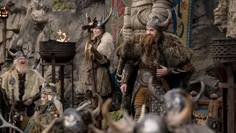 Universal Pictures A man dressed like a viking with a horned helmet, and draped in animal skins. He has a long beard and a woman stands next to him dressed in a similar fashion. People stand around them and a bowl with fire sits in front of them.