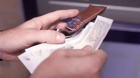 BBC A close-up of hands putting banknotes into a leather wallet