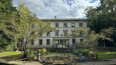 Frannie Brown The front of Fyfield Manor. It's a large white building with pastel green trim. In front of it is a pond with lily pads.