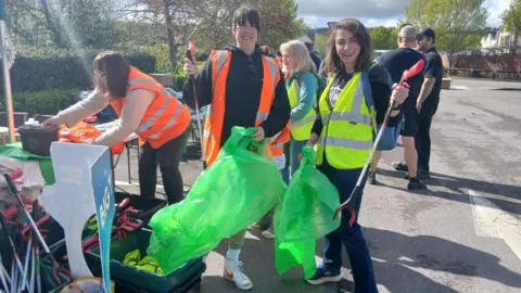 Two women in high-vis vests are standing outside holding rubbish bags and litter pickers. They are smiling at the camera.
