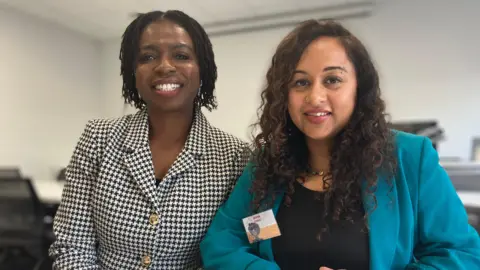 Jon Wright/BBC Dr Yemisi Sloane and Dr Mirna Guha sit next to each other, smiling at the camera. They are in a lecture room.