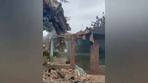 An excavator with a large pneumatic drill attachment breaks up a brick bus shelter. It is working on the roof and a mural on a back wall can just be seen through the dust.