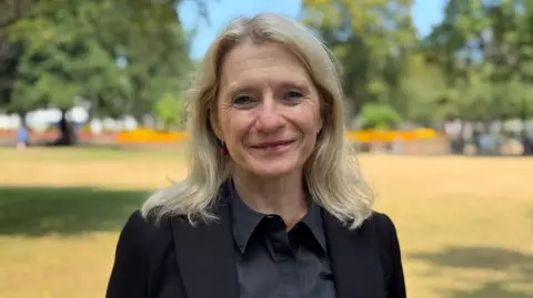 A portrait of woman with shoulder-length light hair wearing a black blazer over a dark buttoned shirt, standing outdoors in a park. The background is softly blurred, showing trees, grass, and flowerbeds in daylight. The person is centered in the frame and looking toward the camera.