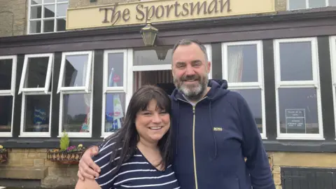 James and Tasmin Kaminiski smile at the camera. James has short brown hair and a greying beard. He is wearing a navy hooded top. Tasmin has ling, dark hair and is wearing a blue and white striped top.