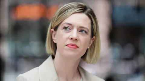 A woman with a blonde bob, wearing a beige blazer and a white top staring off into the distance.