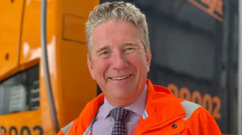 Emma Baugh/BBC Mr Smith is smiling at the camera while wearing an orange high vis jacket. Underneath he is wearing a blue tie with circle patterns and a shirt. He has light blonde/grey hair and is standing in front of the locomotive. Its is blurred into the background but its black and yellow colours can be made out.