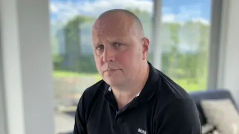 A bald man is looking directly at the camera. He is wearing a black polo shirt with small white writing to the right of his chest which reads 'Boss'. Behind him is a large window, with out of focus greenery. 