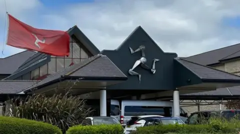 The exterior of Noble's Hospital entrance, you can see the three legs of man and a flag outside, there are well-kept bushes in front.