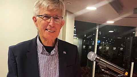 RNLI Clive Rand is wearing a black jacket and a pale shirt underneath it. He is stood behind a podium with an RNLI poster on it.