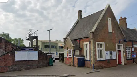 A train station sign attached to a brick wall reads "Totton". Underneath it is a map and a schedule of train times. To the right there is a station building with a red door and a post box in its wall.