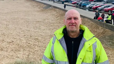 Carl Quartermain is standing on a sandy beach and looking directly at the camera. He is bald, has a grey beard and is wearing a yellow high-vis jacket over a black fleece. On the land, there are dozens of cars in the car park and rows of houses further along the shore.