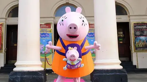 A person in a large pig costume stands outside a theatre with their arms spread wide. A smaller pig in a baby carrier is attached to their chest. The theatre is visible in the background with production posters on the walls.