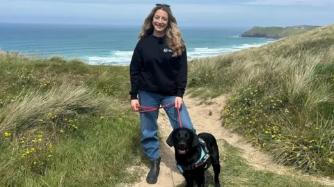 Hannah Barnikel from Cornwall Wildlife Trust is stood on a narrow sandy path through on the sand dunes. She is holding a red dog lead which is attached to a black dog sat in front of her looking at the camera. In the background there is tall grasses and small yellow flowers either side of the path, with the sea in the distance. 
