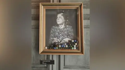 Supplied A framed black and white portrait of a woman looking to the side with marbles flying in the space in front of her and falling in her palms. The bottom of the frame is filled with actual marbles.