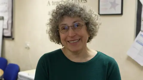 A woman with grey curly hair, wearing round glasses, smiling and wearing a bottle green top