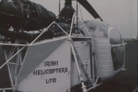 BBC Archive Un pequeño helicóptero enmarcado blanco se ve desde atrás con las palabras 'Irish Helicopters Ltd' impresas en letras negras en un panel trasero. Un motor negro se encuentra por encima del marco con las palas del rotor ligeramente visibles. En el frente hay una pequeña cabina que parece que tendría espacio para solo dos o tres personas. Está hecho de tiras de metal delgadas rodeadas de grandes paneles de ver a través de.  